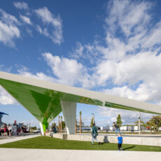 Public park scene with a large green and white angular pavilion, people walking and standing nearby beneath a bright blue sky with scattered clouds.