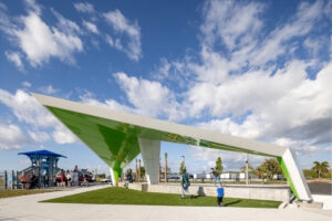 Public park scene with a large green and white angular pavilion, people walking and standing nearby beneath a bright blue sky with scattered clouds.