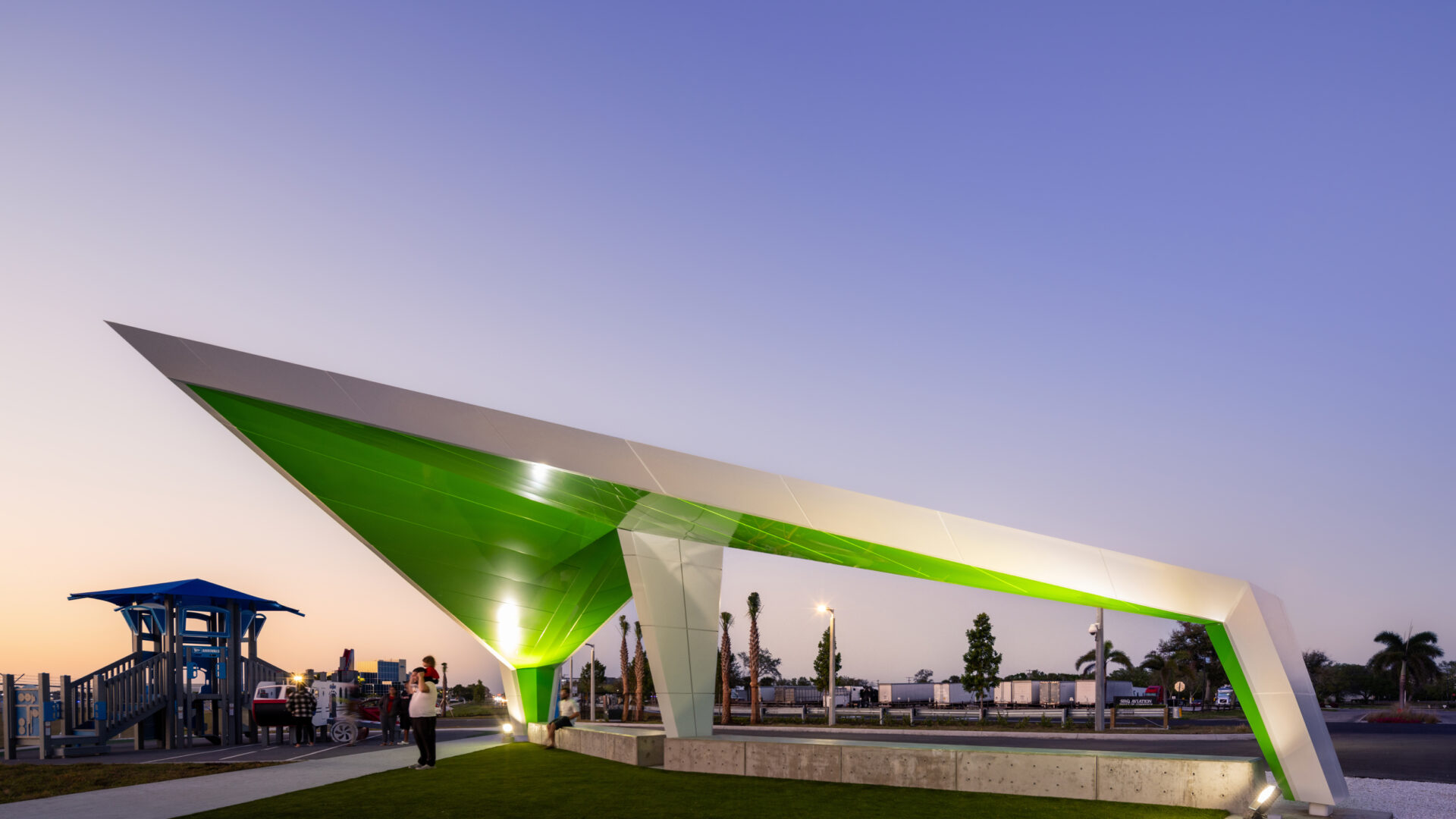 Green and white angular sculpture arching over a plaza at dusk, with people and palm trees in the background.