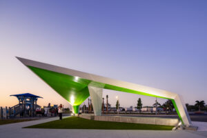 Green and white angular sculpture arching over a plaza at dusk, with people and palm trees in the background.