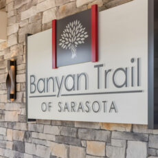 Exterior sign mounted on a stone facade displaying the words 'Banyan Trail' and 'Of Sarasota' with a white tree logo between red decorative posts.