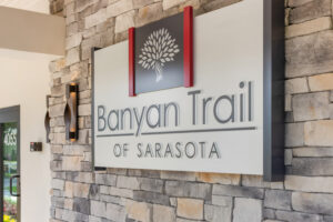 Exterior sign mounted on a stone facade displaying the words 'Banyan Trail' and 'Of Sarasota' with a white tree logo between red decorative posts.