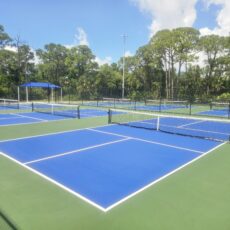 Outdoor tennis facility with multiple blue courts, nets, and surrounding greenery under a blue sky.