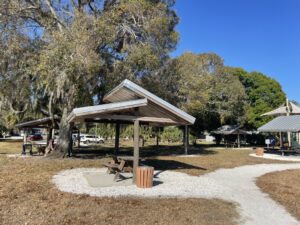 Park scene with a wooden pavilion, picnic table, and tall trees under a clear blue sky.