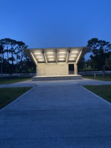 Open-air concrete pavilion with bright overhead lights at dusk, surrounded by trees