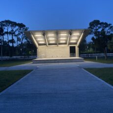Open-air concrete pavilion with bright overhead lights at dusk, surrounded by trees