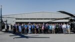 Group of people in business and casual attire posing in a line at an airfield in front of a metal hangar, with helicopters on both sides under a clear blue sky