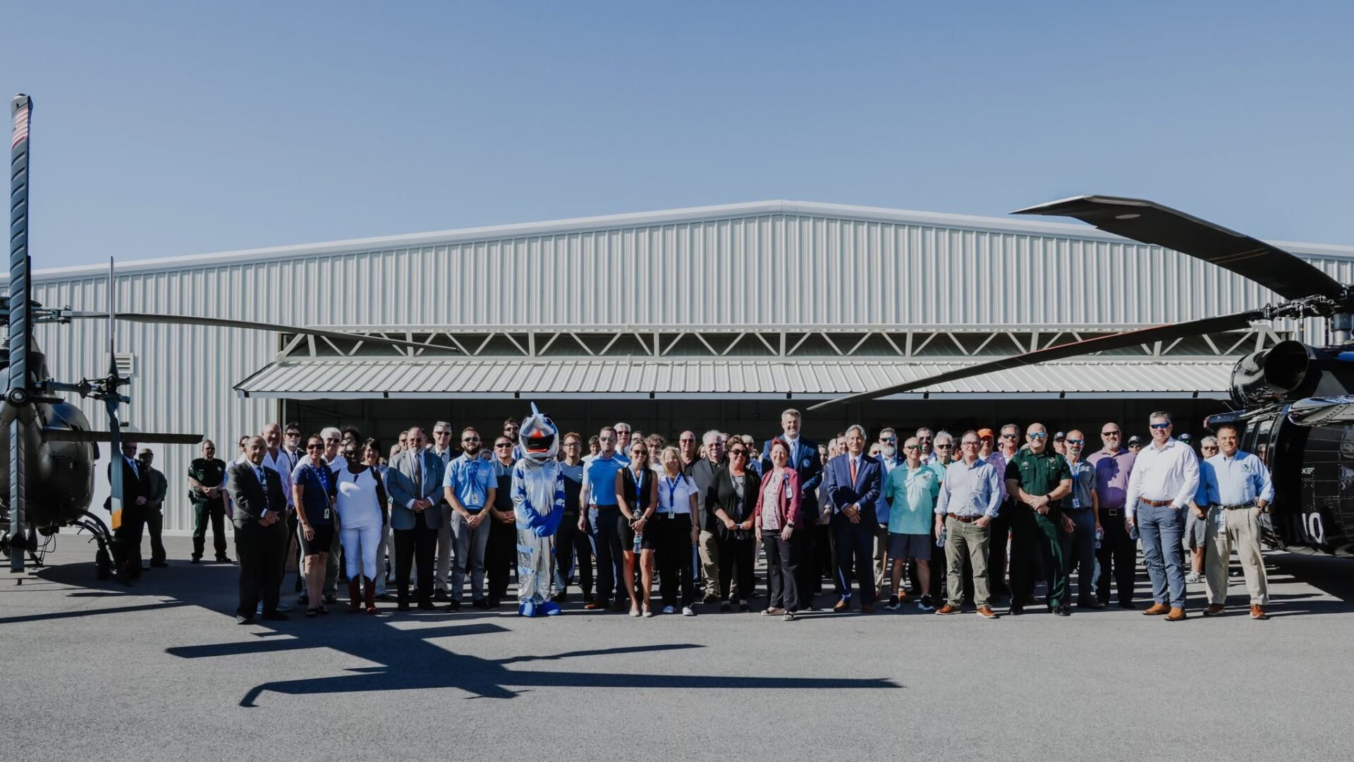 Group of people in business and casual attire posing in a line at an airfield in front of a metal hangar, with helicopters on both sides under a clear blue sky