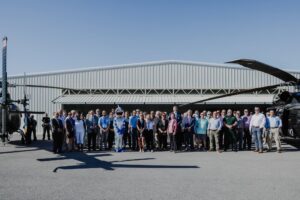 Group of people in business and casual attire posing in a line at an airfield in front of a metal hangar, with helicopters on both sides under a clear blue sky