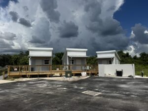 Three small white modular houses on a wooden deck, set on a construction site beneath dramatic storm clouds.